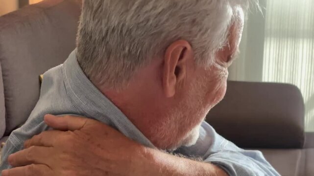 Retired elderly man massaging his aching shoulder while sitting on the couch at home, dealing with back pain, stress, exhaustion, and discomfort due to aging, arthritis, and poor posture