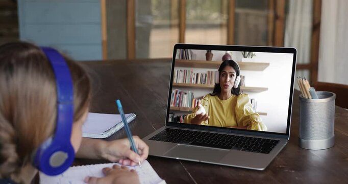 Little girl in headphones focused on writing exercise in copybook during online class with young Indian teacher displayed on laptop screen. Virtual tutoring and distance education, digital technology