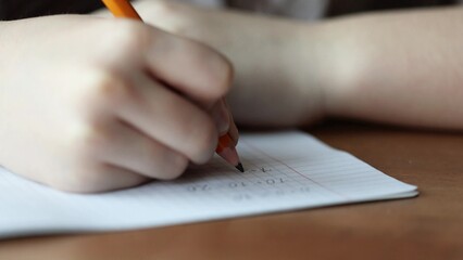 a child writes with a pencil and solves math examples in a checkered notebook. primary school...