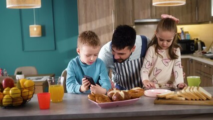 Two little kids brother and sister playing games or watching something on smartphones at the table while their dad prepares breakfast for them in the kitchen - Powered by Adobe