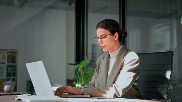 Young professional it specialist latin hispanic business lady working on laptop pc sitting at desk in modern office space. 30s middle eastern indian woman using computer technology app for work online