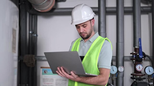 Serious male engineer in hardhat and uniform checks readings on laptop and inspects heating system equipment in boiler room.