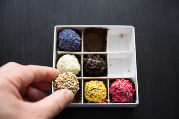A hand delicately holds a nut-covered truffle from a box of assorted colorful truffles. The box contains various truffles with different coatings, including blue, white, yellow, and pink.