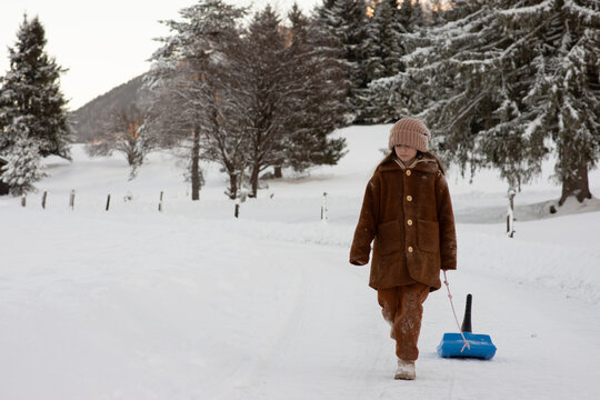 A little girl with a sleigh walks through an alpine village in winter. Snowy landscape and a girl in a brown fur coat and mittens. Winter fun and active recreation with children