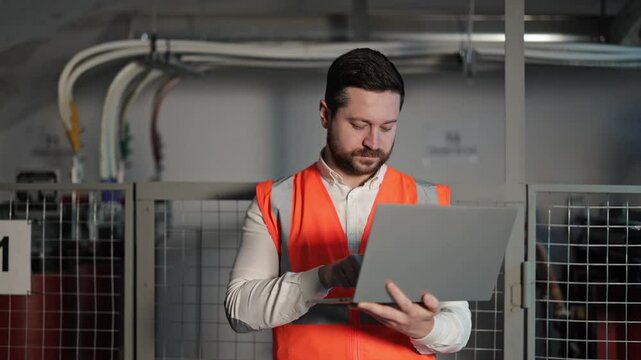 Serious electrical engineer with a beard and wearing an orange safety vest uses laptop next to an electrical control panel in industrial environment. Industrial room with electric devices and wires.
