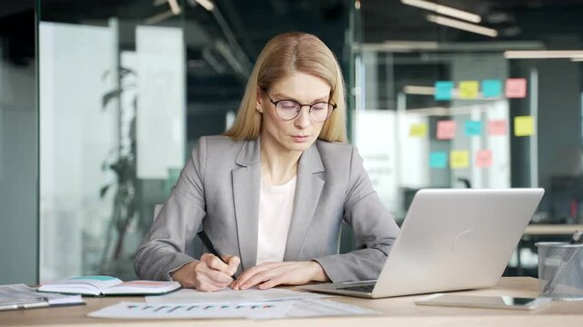 Busy businesswoman is doing paperwork using a laptop while at workplace in business office. Confident accountant working fills out documents, writing, making a tax return or engaged in accounting