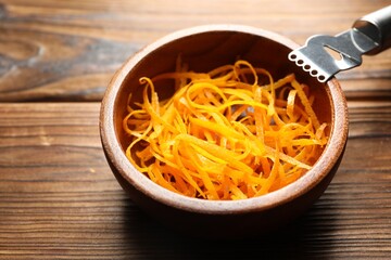 Fresh orange zest in bowl and zester tool on wooden table, closeup