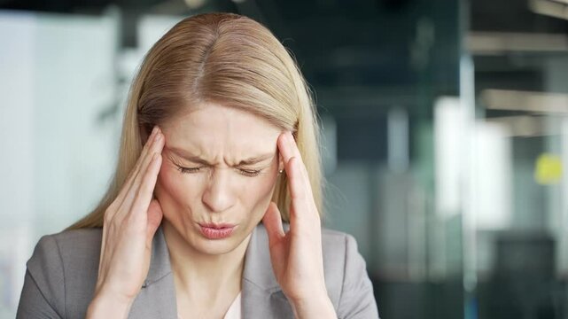 Tired businesswoman in suit suffering from headache while sitting at workplace in business office. Upset blonde female employee feels pain, has migraine, massaging her temples with her hands. Close up