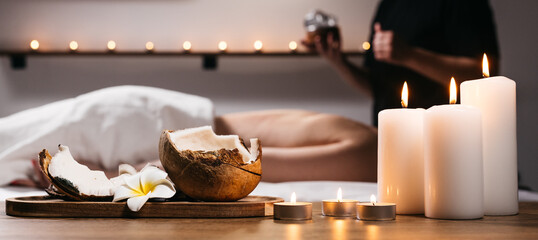 Close-up of a coconut with a tropical flower, candles, and incense in a modern spa. In the background, a body scrub massage is being performed on a young woman. Spa, relaxation, and body care concept.