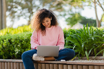 pretty curly young woman sitting in park working on laptop remote job