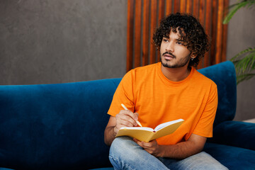 Young Indian man wear orange casual clothes sits on blue sofa couch write down memories in notebook...