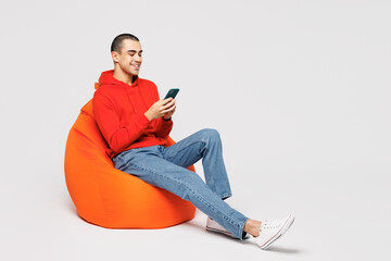 Full body young middle eastern man he wearing red hoody casual clothes sit in bag chair hold in hand use mobile cell phone isolated on plain solid white background studio portrait. Lifestyle concept.