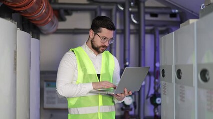 Serious, focused male technician in uniform types on laptop near heating system equipment in boiler room. Man works with modern technologies.