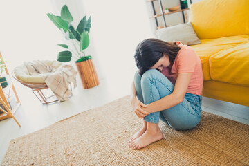 Young woman sitting on the floor looking thoughtful and introspective in a cozy living room with...