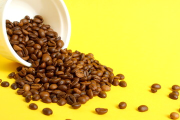 A white ceramic bowl with roasted coffee beans is overturned on a yellow background.
