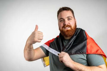 Young man with a red beard draped in the German flag, giving a thumbs up in a studio with a neutral...