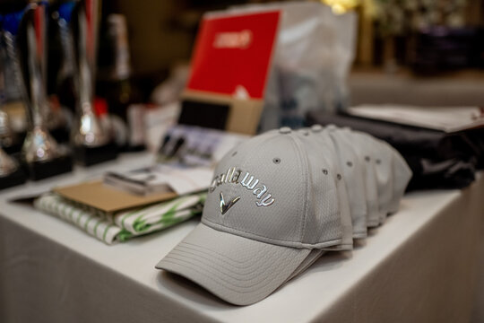 A row of gray branded caps neatly arranged on a table with trophies, cloth, and gift items in the background, in a professional setting.