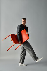 A young man dressed stylishly walks with a red chair held up in a modern studio.