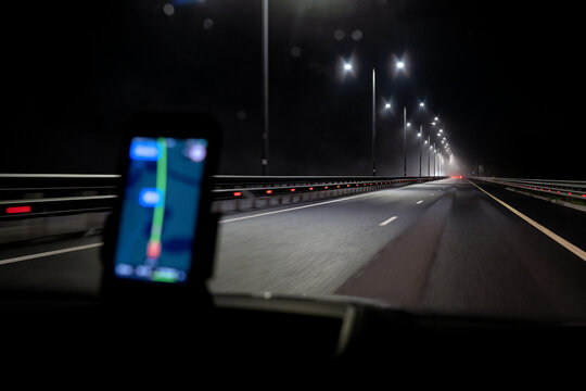 Dark night highway goes far ahead, red lights of cars are visible. There is a blurry smartphone with an open navigation app. Background. - Powered by Adobe