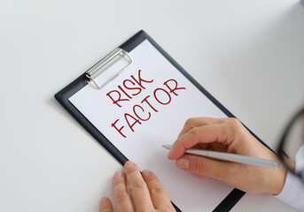 A person is writing on a clipboard with the word Risk Factor written in red. The clipboard is on a table and the person is using a pen to write