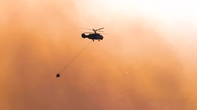 A helicopter flies over a forest carrying a water bucket to combat a wildfire. The hazy sky and rising smoke highlight the intensity of the fire and the ongoing efforts to bring it under control.