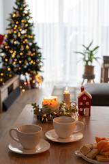 Cozy Christmas scene with two empty white cups on wooden table, festive candles, and plate of snacks in the foreground, with decorated Christmas tree glowing in soft bokeh lights in the background.