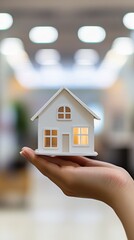 Businesswoman is holding a small white house model in her hand, symbolizing investment, real estate, and homeownership, with a blurred office background