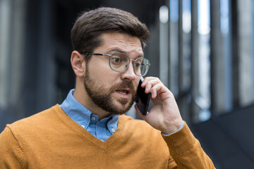 Close-up photo of a sad young man in glasses standing on the street and worriedly talking on the phone