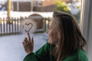 Beautiful woman painting  a heart on a misty window with her finger. Love,  valentines day celebration concept