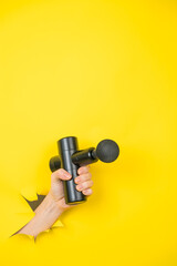 Female hand sticking out with massage gun from yellow paper background. Vertical photo. 