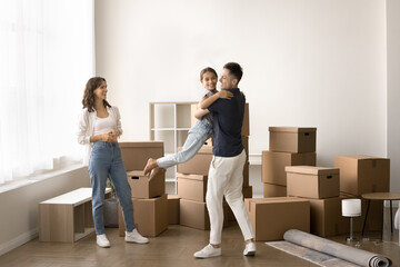 Happy family celebrate relocation to new house, full-length shot. Young daddy lifting, spinning his little daughter stand together with wife in empty living room with heap of stacked cardboard boxes
