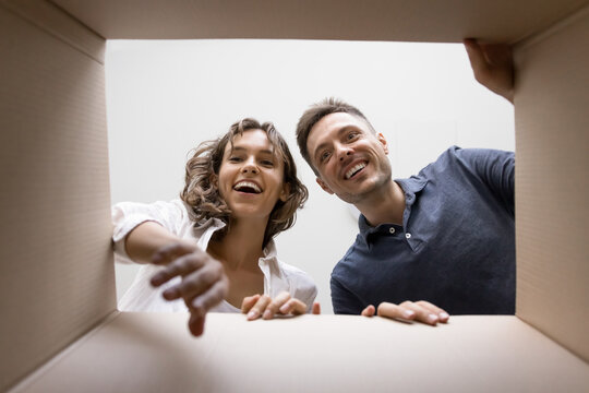 Young amazed couple opening cardboard box, enjoy received order, bought goods via electronic commerce website. Marketplace app clients take out things, box bottom view. Delivery services, e-shopping - Powered by Adobe
