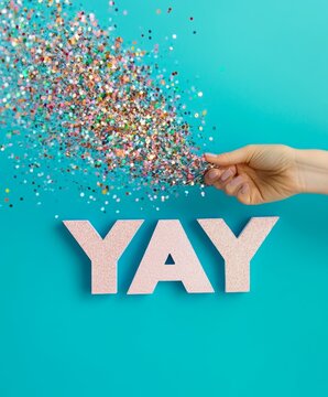 hand throwing colorful confetti on pink sign YAY against blue background