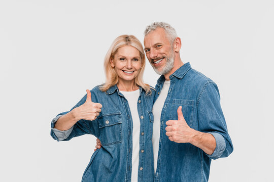 Excited cheerful funny couple two friends elderly gray-haired man blonde woman wearing denim shirt casual clothes standing showing thumbs up signs gesture isolated on white background studio portrait