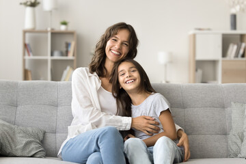 Portrait of young mother cuddling her little 6s daughter seated on sofa smiling at camera, enjoy priceless carefree time together at home, feeling unconditional love. Happy Mothers Day, custody, ties