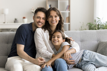 Obraz premium Portrait of happy Latin parents enjoy sweet family moments with adorable little daughter, sit on couch at home, looking at camera with positive expression, exuding warmth, joy and unconditional love