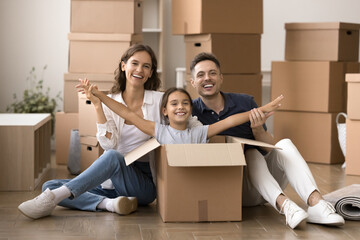 Cute little girl sitting inside big cardboard box, smile looking at camera, posing with loving parents at their new house on move-out or relocation day, celebrate first dwelling purchase, feel happy