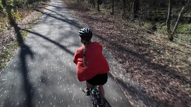 Mature woman wearing a red shirt and helmet bikes down a paved path through a forest, surrounded by fallen leaves and trees on either side on C&O Canal.