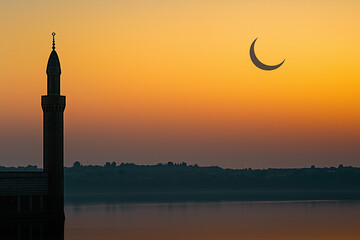 Fototapeta premium Minaret silhouette at sunset with crescent moon illuminating orange sky over river