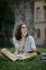 A male student sits on the grass in a park outdoors, smoking a cigarette while holding a notebook....