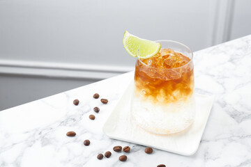 Refreshing espresso tonic drink with slice of lime and coffee beans on white marble table