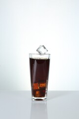 Ice cube falling into glass with tasty cola on mirror table against white background