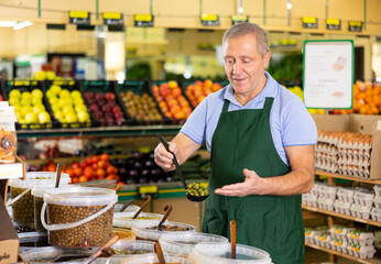 Portrait of smiling elderly salesman in apron arranging various types of pickled olives in hypermarket