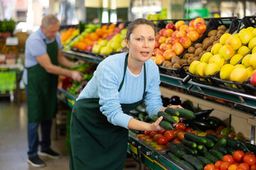 Portrait of smiling busy female merchandiser with cucumber in hands at grocery supermarket