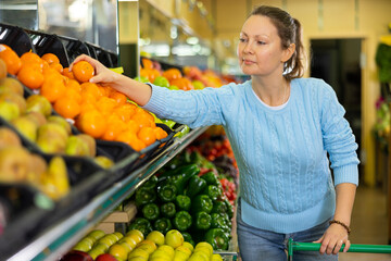 Pleased middle-aged woman buyer purchasing fresh mandarins at the counter in large vegetable shop