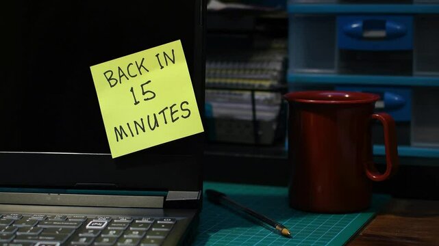Office setting man&rsquo;s hand sticking yellow Back in 15 minutes note message to laptop mood lighting copy space