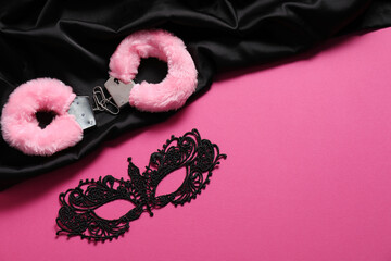 Fluffy handcuffs, lace mask and black fabric on pink background, flat lay. Space for text