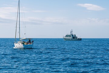 A small sailboat with three individuals is being inspected by an armed forces vessel in the open sea, showcasing maritime security operations and the importance of safeguarding waters.