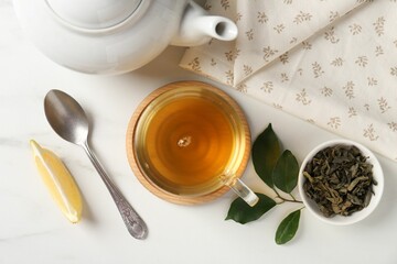 Refreshing green tea in cup, spoon, leaves and slice of lemon on white table, flat lay