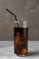 Refreshing cola with ice cubes and drinking straw in glass on light textured table against grey background, closeup